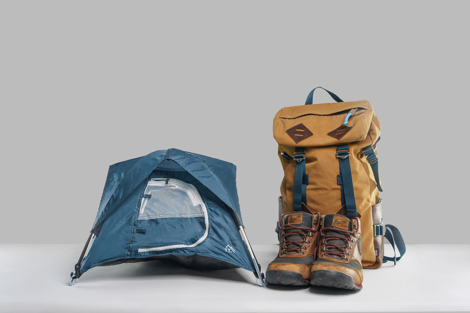 Blue Tiny Tent sitting next to hiking boots and a rucksack on gray background