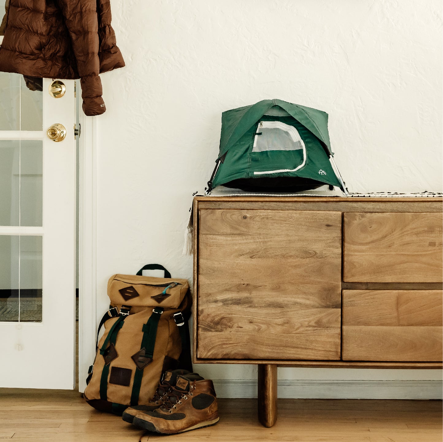 Green tiny tent shown up in the entryway of a home. There is a backpack and hiking shoes in the image.