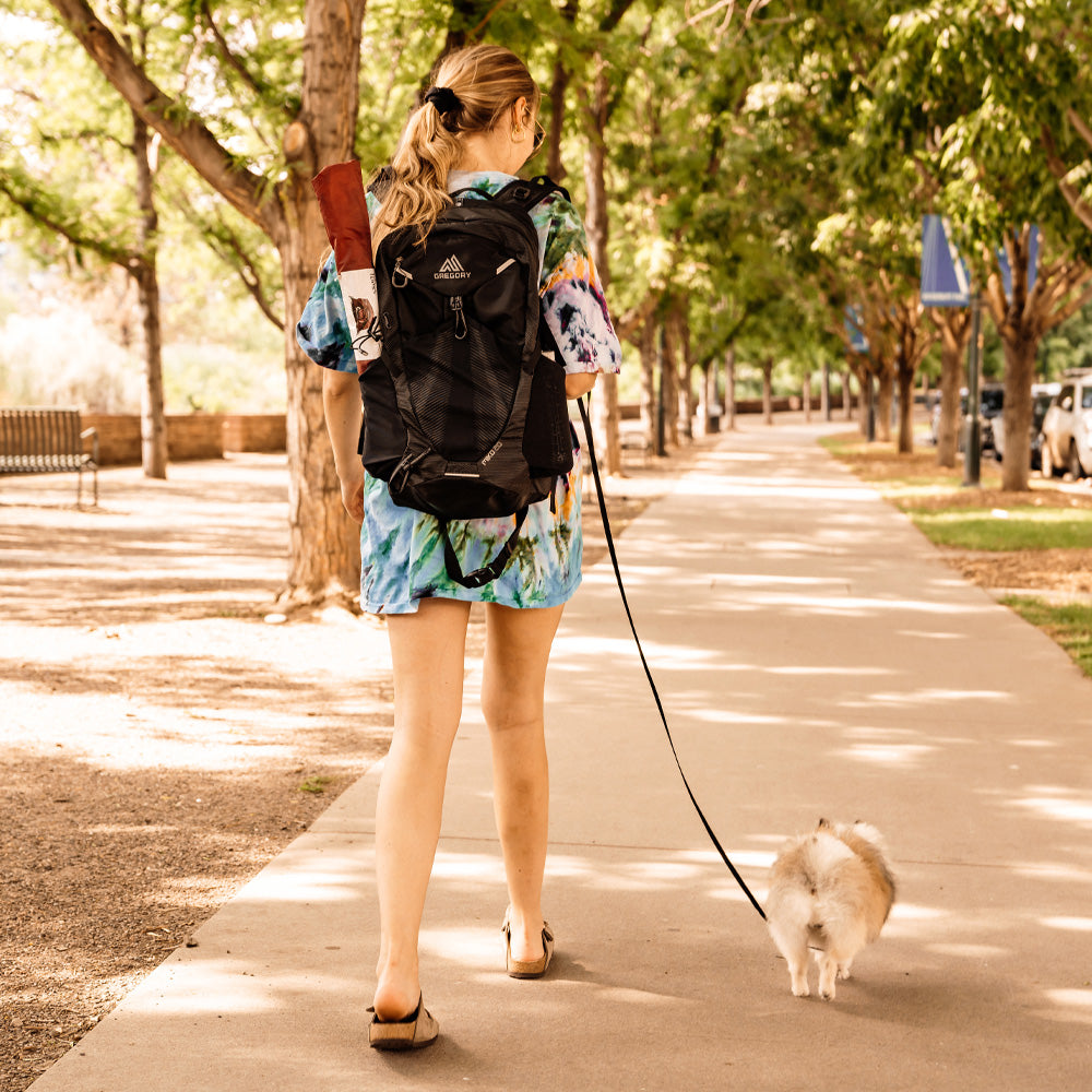 woman walking down shady sidewalk with her small fluffy dog, a packed up berry tiny tent in her backpack