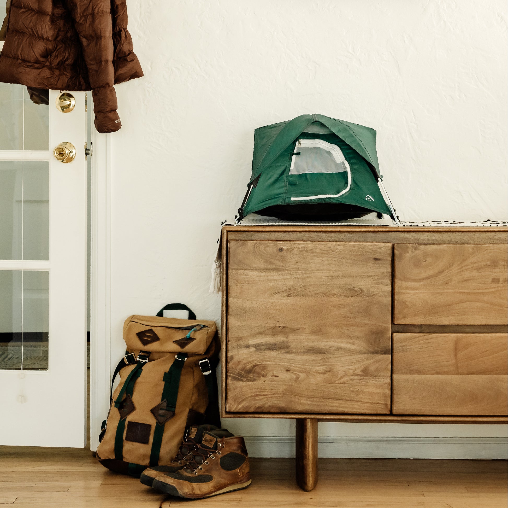Green tiny tent shown up in the entryway of a home.  There is a backpack and hiking shoes in the image.